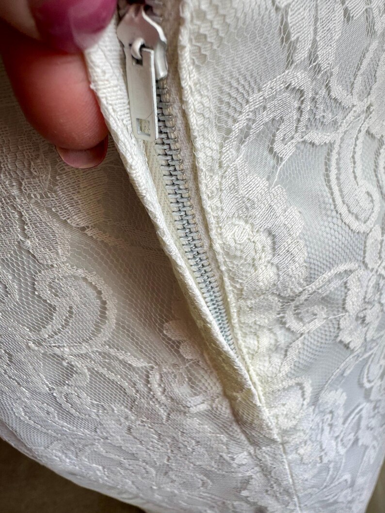 May include: Close-up of a white lace fabric with a zipper. The lace features a floral pattern. The zipper is white and partially open, revealing the fabric's texture and construction. The overall aesthetic is delicate and elegant.