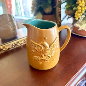 May include: A tan ceramic pitcher with a teal interior and a handle. The pitcher features a raised relief of an eagle with stars. The pitcher is sitting on a dark brown surface.