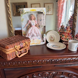 May include: A display featuring a Barbie doll in a pink dress, a wicker basket, and Peter Rabbit themed dishware. The doll is in a collector's box. A decorative mirror with a gold frame is in the background.