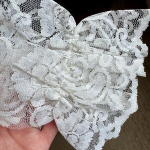 May include: Close-up of white lace fabric with a floral pattern. The delicate lace is gathered and layered, creating a textured effect. The intricate design and soft color suggest a vintage or romantic aesthetic, suitable for apparel or craft projects.