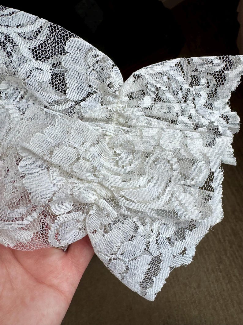 May include: Close-up of white lace fabric with a floral pattern. The delicate lace is gathered and layered, creating a textured effect. The intricate design and soft color suggest a vintage or romantic aesthetic, suitable for apparel or craft projects.