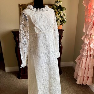 May include: A vintage white lace dress with long sleeves and a ruffled neckline. The dress is displayed on a black mannequin. The dress has a floral lace pattern and a fitted bodice. The dress is floor-length.