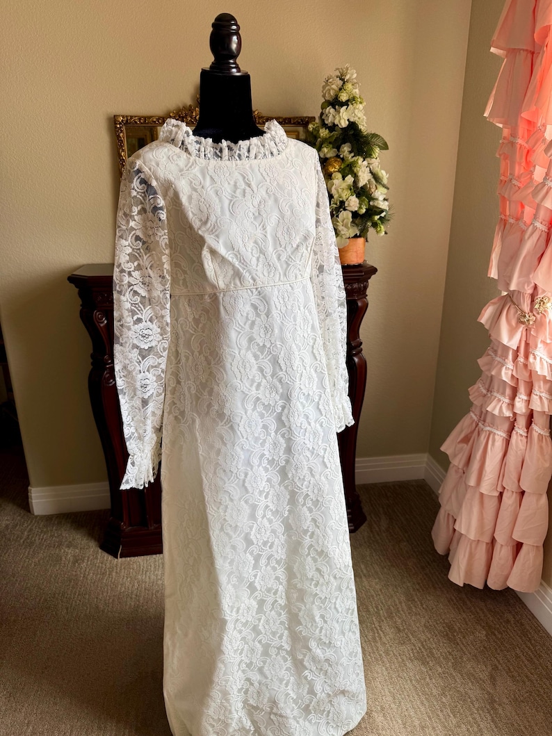 May include: A vintage white lace dress with long sleeves and a ruffled neckline. The dress is displayed on a black mannequin. The dress has a floral lace pattern and a fitted bodice. The dress is floor-length.