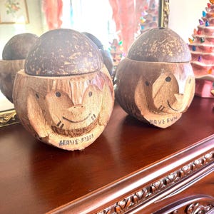 May include: Two carved wooden containers with lids. The containers are brown with a carved face design and the words "HAVE FUN" carved below the face. The lids are a darker brown. The containers are sitting on a dark brown wooden surface.