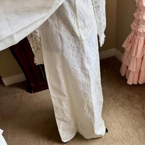 May include: A white lace wedding dress with long sleeves and a flowing skirt. The dress features intricate floral lace detailing and a high neckline. The dress is floor-length and appears to be vintage or antique.