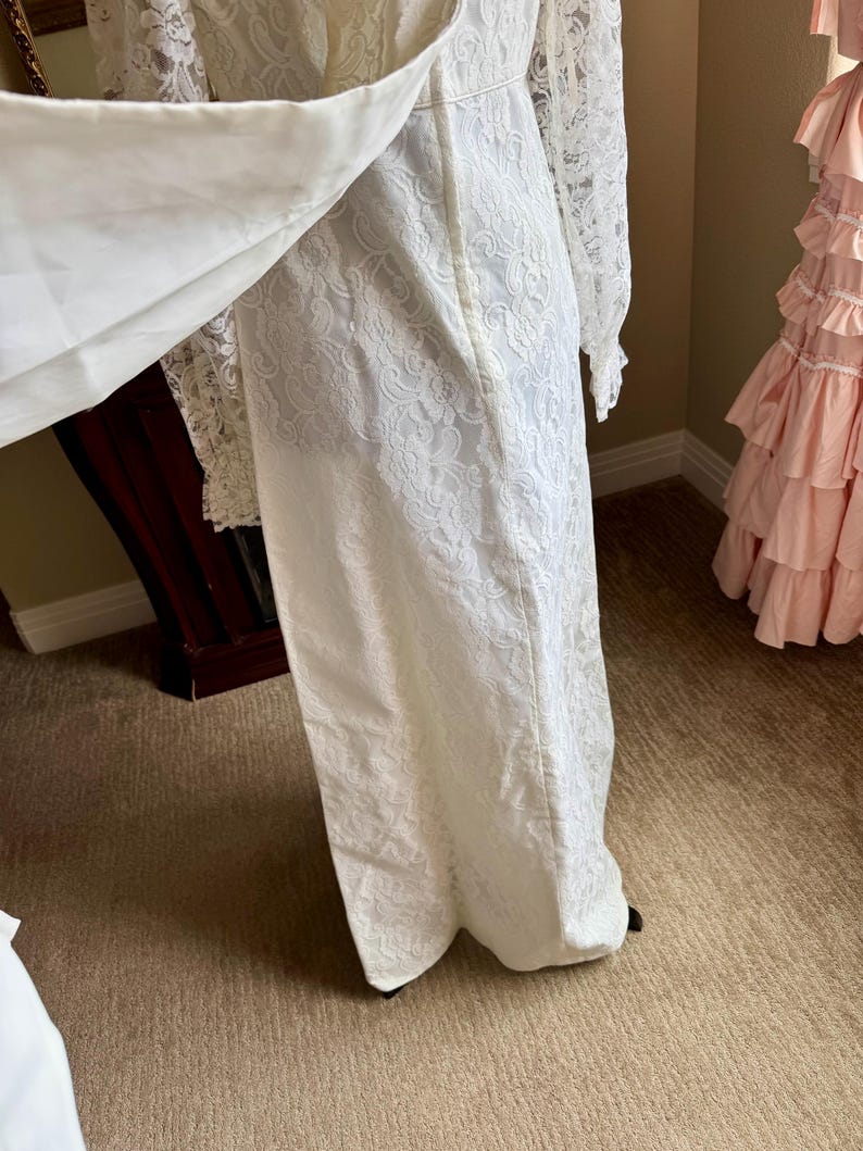 May include: A white lace wedding dress with long sleeves and a flowing skirt. The dress features intricate floral lace detailing and a high neckline. The dress is floor-length and appears to be vintage or antique.
