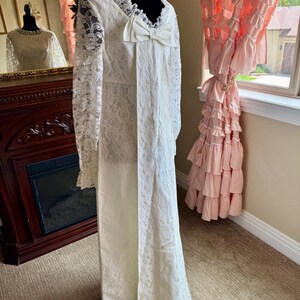 May include: A vintage ivory wedding dress with long lace sleeves and a large bow. The dress features intricate lace detailing and a high neckline. The dress is displayed on a black mannequin. The dress is floor length.
