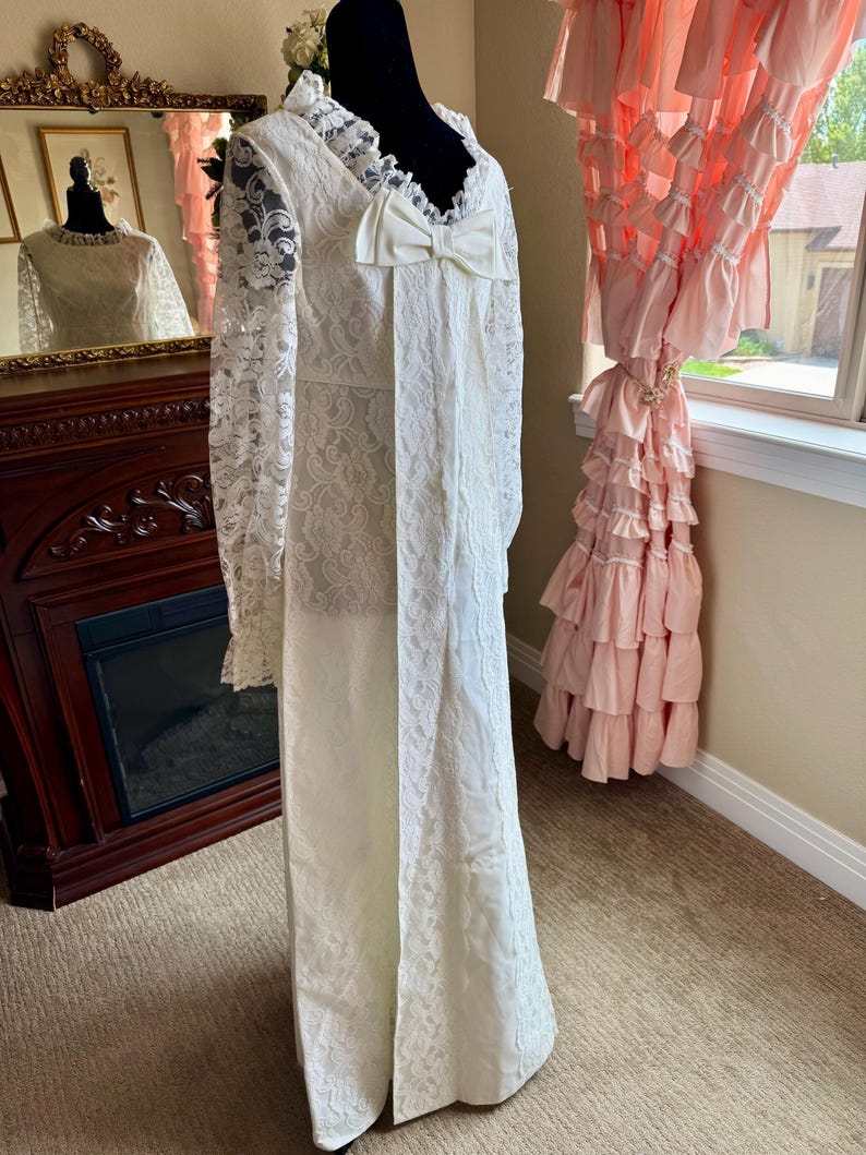 May include: A vintage ivory wedding dress with long lace sleeves and a large bow. The dress features intricate lace detailing and a high neckline. The dress is displayed on a black mannequin. The dress is floor length.