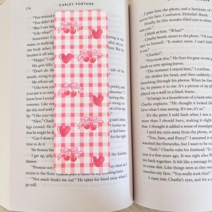 May include: A pink and white gingham bookmark with repeating patterns of cherries, bows, and hearts. The bookmark is inserted into a book with text on the pages.