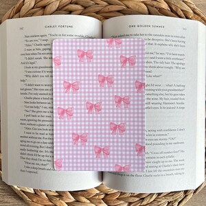 May include: A pink and white gingham bookmark featuring pink bow illustrations. The bookmark is rectangular and is positioned between the pages of an open book. The book is open to pages 162 and 163.