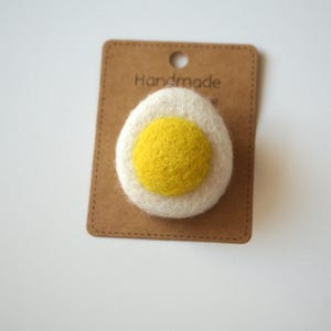 May include: A handmade felt egg pin with a white yolk and a yellow yolk, attached to a brown card. The card has a small hole at the top and the word "Handmade" printed on it. The pin is a fun accessory.