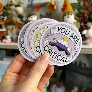 Op de afbeelding: Een stapel ronde stickers met een lavendelkleurige achtergrond en de woorden "YOU ARE CRITICAL" in het wit. De stickers tonen een non-binaire pride-vlag en dobbelstenen. De stickers worden in een hand gehouden.