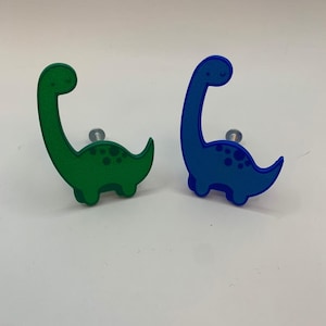Könnte beinhalten: Zwei dinosaurierförmige Schrankknöpfe, einer grün und einer blau. Die Knöpfe haben ein cartoonartiges Design und sind perfekt für ein Kinderzimmer.