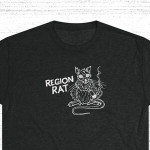 Op de afbeelding: Donkergrijs t-shirt met een witte afbeelding van een sigaret rokende rat, met de tekst "REGION RAT" erboven. De rat is getekend in een gedetailleerde, cartoonachtige stijl. Het t-shirt is gemaakt van een zachte, getextureerde stof.