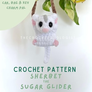 Puede incluir: Un llavero de coche, bolso y llave de crochet blanco de una ardilla voladora, con detalles en rosa y ojos negros. El amuleto tiene una correa marrón y cuelga frente a una planta. El texto en la imagen dice "CAR, BAG & KEY CHARM PAL" y "CROCHET PATTERN SHERBET THE SUGAR GLIDER."