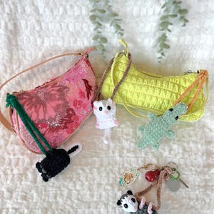 May include: Two shoulder bags, one pink floral and one yellow quilted, are shown with handmade crochet keychains. The keychains feature a skunk, a white cat, a green alligator, and a grey opossum.