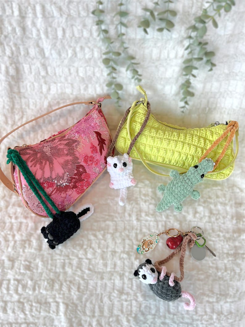 May include: Two shoulder bags, one pink floral and one yellow quilted, are shown with handmade crochet keychains. The keychains feature a skunk, a white cat, a green alligator, and a grey opossum.