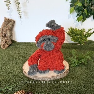 May include: A handmade, plush orangutan toy in shades of red and gray, sitting on a wooden slice. The crochet animal has a textured, fuzzy appearance. The text "THE CROCHET ZOOLOGIST" is visible at the bottom. The toy is displayed on a green surface.