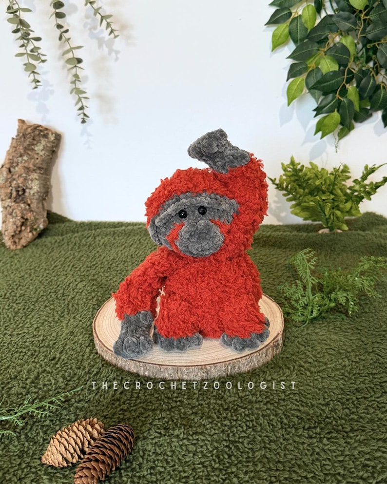 May include: A handmade, plush orangutan toy in shades of red and gray, sitting on a wooden slice. The crochet animal has a textured, fuzzy appearance. The text "THE CROCHET ZOOLOGIST" is visible at the bottom. The toy is displayed on a green surface.