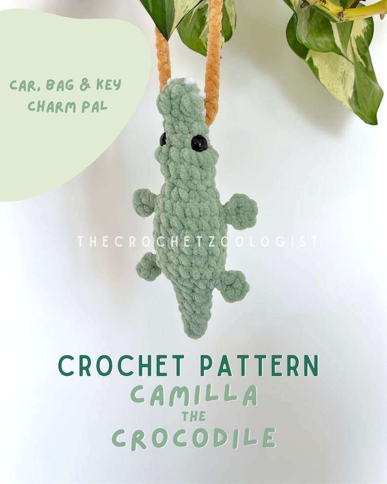 May include: A crocheted green crocodile charm with black eyes and small limbs, suspended by a tan cord. The image includes text: "CAR, BAG & KEY CHARM PAL" and "CROCHET PATTERN CAMILLA THE CROCODILE". The Crochet Zoologist is also printed on the image.