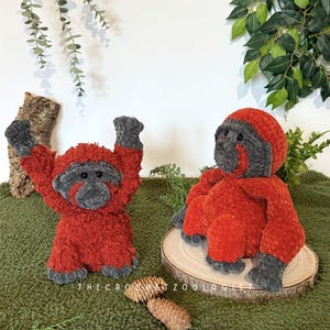 May include: Two plush orangutan toys. One is standing with arms raised, the other is sitting. Both are red with gray faces, hands, and feet. The text "THE CROCHET ZOOOLOGIST" is visible at the bottom. The toys are on a green surface.