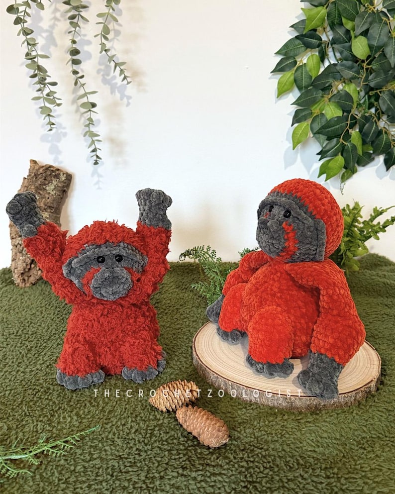 May include: Two plush orangutan toys. One is standing with arms raised, the other is sitting. Both are red with gray faces, hands, and feet. The text "THE CROCHET ZOOOLOGIST" is visible at the bottom. The toys are on a green surface.