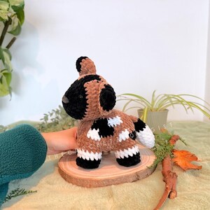 May include: A handmade crochet dog toy in brown, black, and white. The dog has a black head and ears, a brown body with black and white spots, and white feet. It is displayed on a wooden slice.