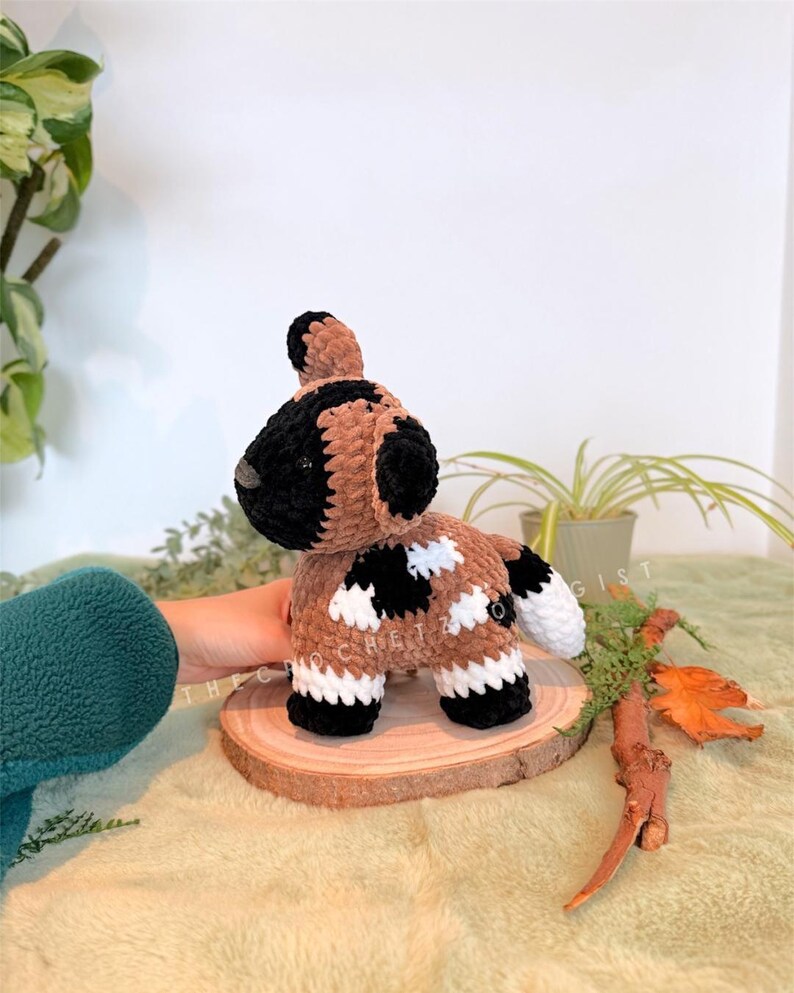 May include: A handmade crochet dog toy in brown, black, and white. The dog has a black head and ears, a brown body with black and white spots, and white feet. It is displayed on a wooden slice.