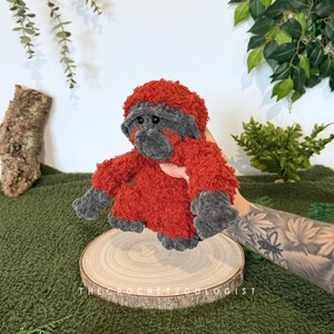 May include: A handmade crochet orangutan plush toy. The plush has a red body and head with gray accents on the face, hands, and feet. The toy is displayed on a wooden slice with the text "THE CROCHET ZOOOLOGIST".