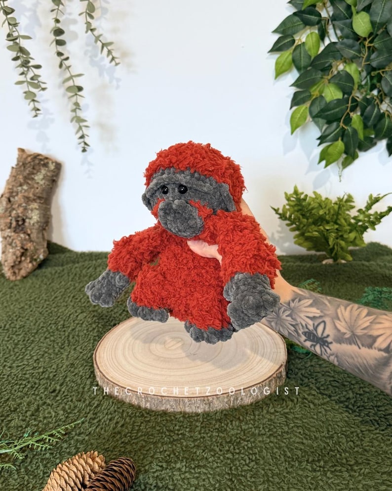May include: A handmade crochet orangutan plush toy. The plush has a red body and head with gray accents on the face, hands, and feet. The toy is displayed on a wooden slice with the text "THE CROCHET ZOOOLOGIST".