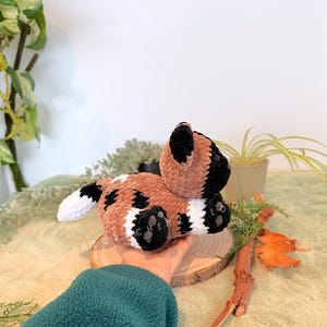 May include: A handmade crochet dog plushie in brown, black, and white. The plush toy is lying on a wooden slice, with the text "THE CRECHETIST" visible. The dog has black paws and ears, with white accents.