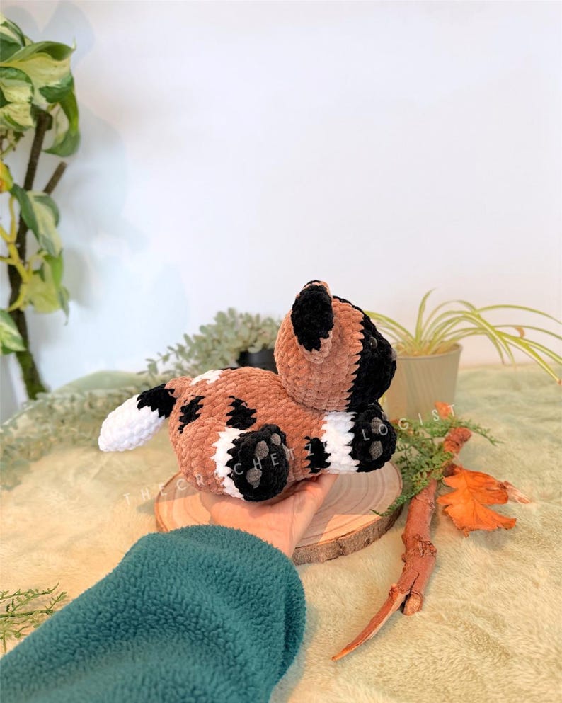 May include: A handmade crochet dog plushie in brown, black, and white. The plush toy is lying on a wooden slice, with the text "THE CRECHETIST" visible. The dog has black paws and ears, with white accents.
