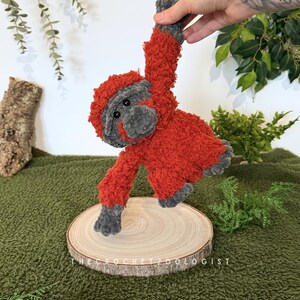 May include: A handmade, plush orangutan doll in shades of red and gray. The crochet animal is suspended above a wooden slice, with the text "THE CROCHET ZOOLOGIST" visible. The doll has a fuzzy texture and is set against a green background.