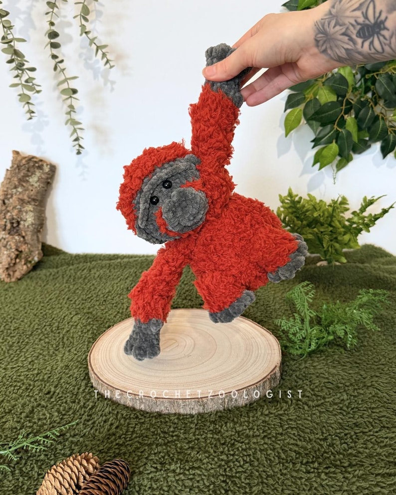 May include: A handmade, plush orangutan doll in shades of red and gray. The crochet animal is suspended above a wooden slice, with the text "THE CROCHET ZOOLOGIST" visible. The doll has a fuzzy texture and is set against a green background.