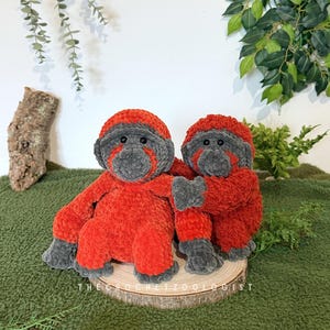 May include: Two handmade crochet orangutan plush toys. The plush toys are red and gray, with the text "THE CROCHET ZOOOLOGIST" on the wooden base. The orangutans are sitting on a wooden slice, with a green background.