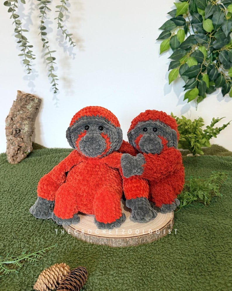 May include: Two handmade crochet orangutan plush toys. The plush toys are red and gray, with the text "THE CROCHET ZOOOLOGIST" on the wooden base. The orangutans are sitting on a wooden slice, with a green background.