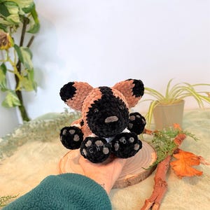 May include: A handmade crochet African wild dog plush toy. The plush features a black and tan color scheme with black paws and ears. The toy is sitting on a wooden slice, with the text "@crochetzoo" visible on the paws.