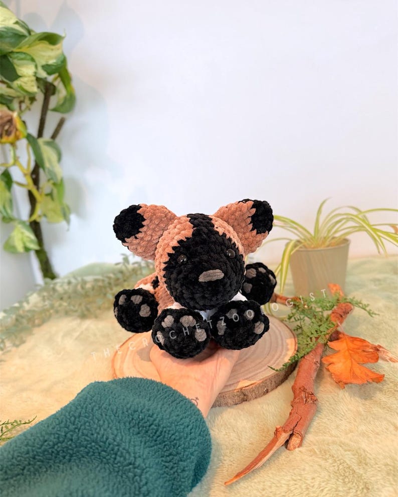 May include: A handmade crochet African wild dog plush toy. The plush features a black and tan color scheme with black paws and ears. The toy is sitting on a wooden slice, with the text "@crochetzoo" visible on the paws.
