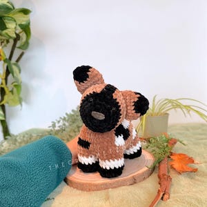 May include: A handmade crochet African wild dog plushie. The plushie features a brown body with black and white markings, black ears, and a black muzzle. The plushie is sitting on a wooden slice. The text "THE CROCHET ZOO" is visible.