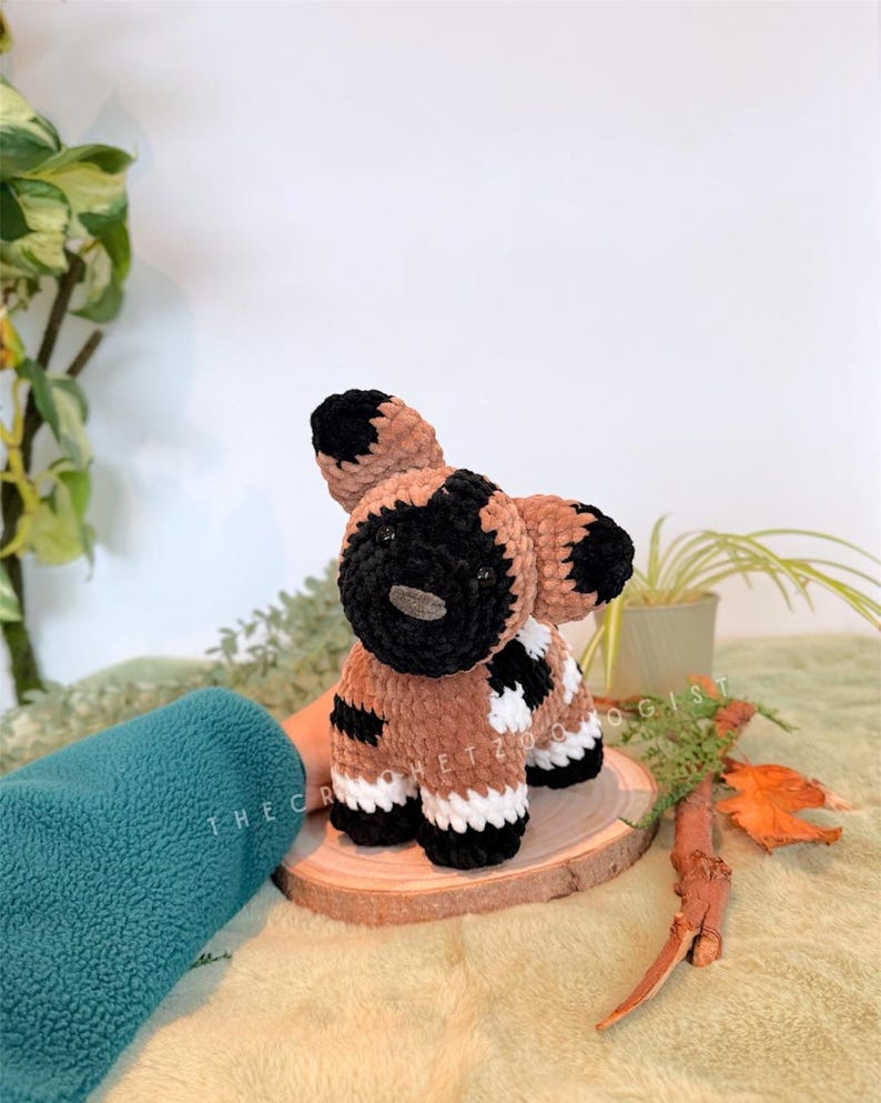 May include: A handmade crochet African wild dog plushie. The plushie features a brown body with black and white markings, black ears, and a black muzzle. The plushie is sitting on a wooden slice. The text "THE CROCHET ZOO" is visible.