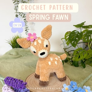 May include: A crocheted fawn with a pink flower on its head, white spots, and brown hooves. The text "CROCHET PATTERN SPRING FAWN" is displayed above the fawn. The fawn is on a wooden slice with purple and blue crocheted flowers.