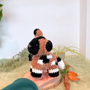 May include: A handmade crochet dog toy in brown, black, and white. The dog has a black head and ears, a brown body with black and white patches, and white paws. The toy is sitting on a wooden slice.