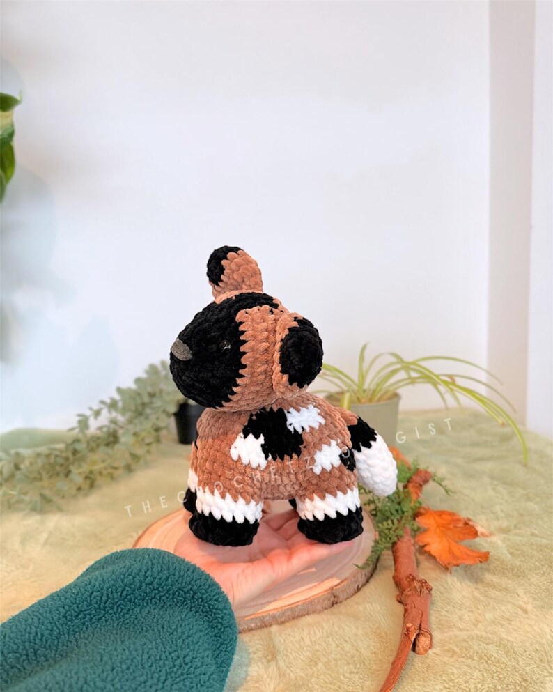 May include: A handmade crochet dog toy in brown, black, and white. The dog has a black head and ears, a brown body with black and white patches, and white paws. The toy is sitting on a wooden slice.