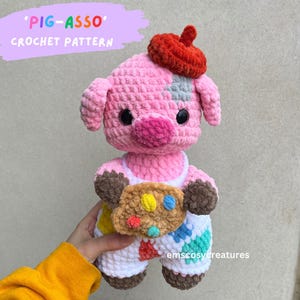 May include: A pink crocheted pig wearing a red beret and holding a brown crocheted paint palette. The pig is wearing a white and colorful crocheted outfit. The text 'Pig-Asso' and 'Crochet Pattern' is visible in the image.