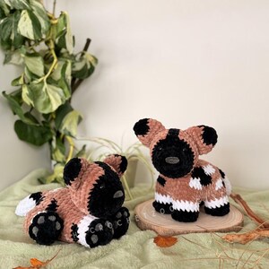 May include: Two handmade crochet African wild dog plushies. One is lying down, the other is upright. Both have brown, black, and white patterns. The plushies are on a green surface with a wooden slice and foliage in the background.