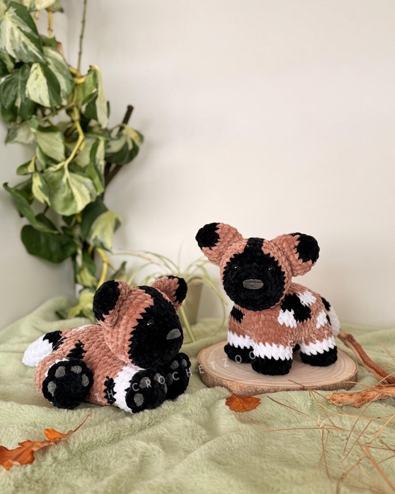 May include: Two handmade crochet African wild dog plushies. One is lying down, the other is upright. Both have brown, black, and white patterns. The plushies are on a green surface with a wooden slice and foliage in the background.