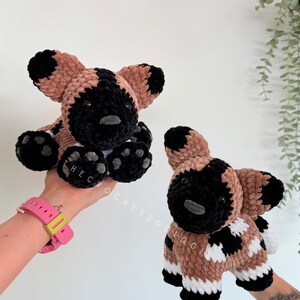 May include: Two handmade crochet African wild dog plushies. The plushies have a black and tan color scheme with black paws and ears. The text "THEC" is visible on one paw. The other plushie has the text "OCHETZOO QG" on its side.