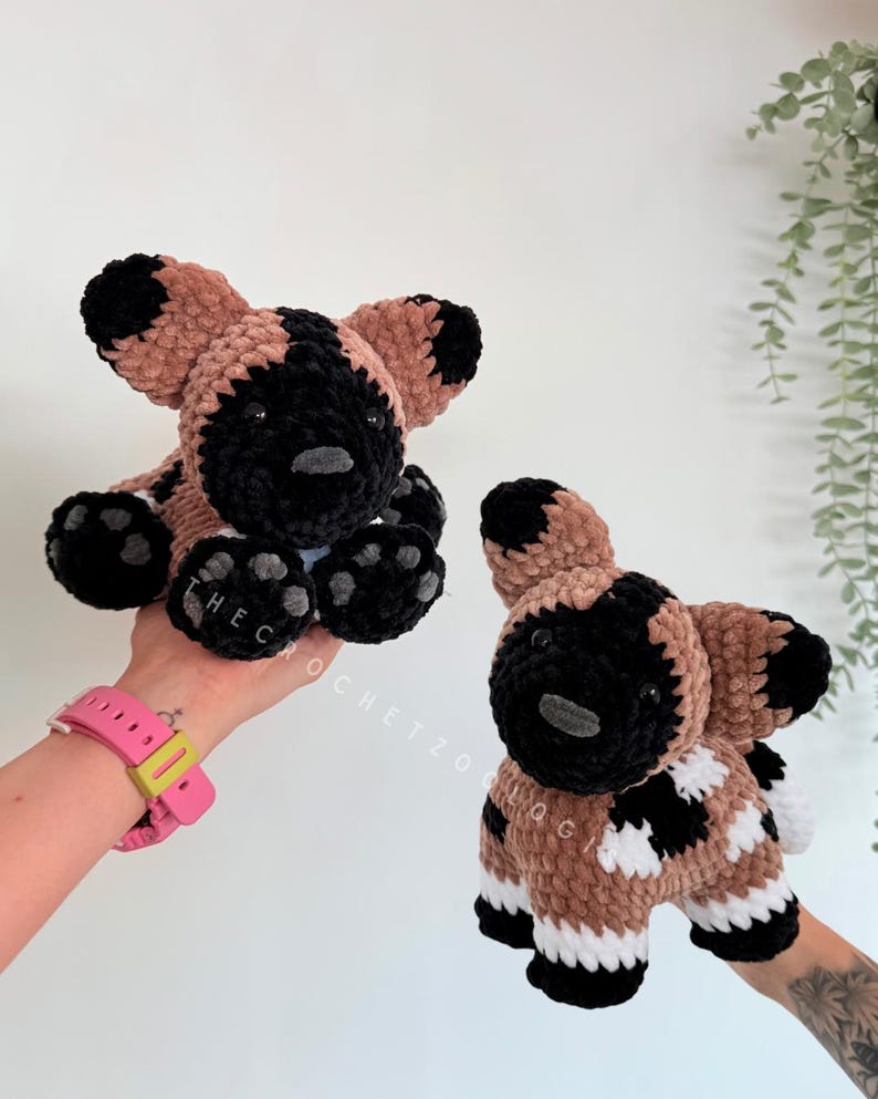 May include: Two handmade crochet African wild dog plushies. The plushies have a black and tan color scheme with black paws and ears. The text "THEC" is visible on one paw. The other plushie has the text "OCHETZOO QG" on its side.