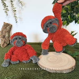 May include: Two plush orangutan toys. One is seated, the other is hanging. Both are orange with gray faces, hands, and feet. They are on a green surface with a wooden slice and the text "THECROCHETZOOOLOGIST".