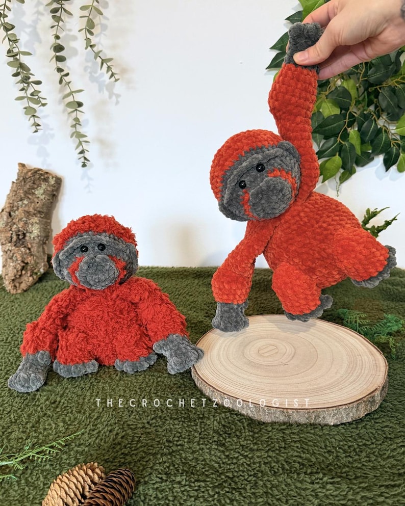 May include: Two plush orangutan toys. One is seated, the other is hanging. Both are orange with gray faces, hands, and feet. They are on a green surface with a wooden slice and the text "THECROCHETZOOOLOGIST".